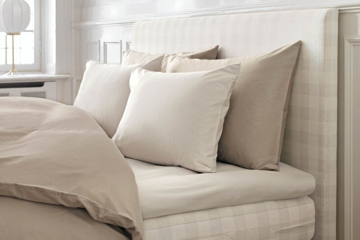 A brightly lit bedroom featuring a bed with neutral beige linens and a striped headboard under natural window light.