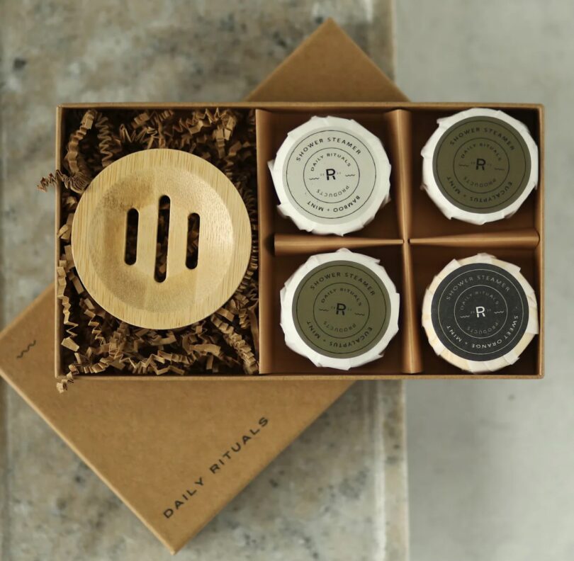 An open gift box containing several round shower steamer tablets next to a bamboo holder.