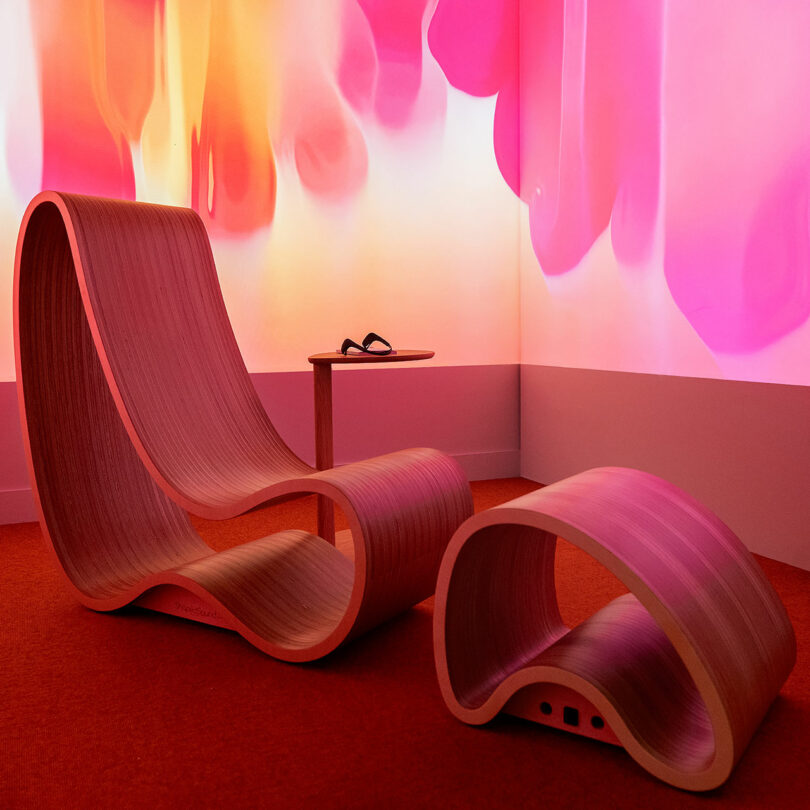 A curved wooden lounge chair and footrest on a red carpet with pink abstract light patterns on the wall.