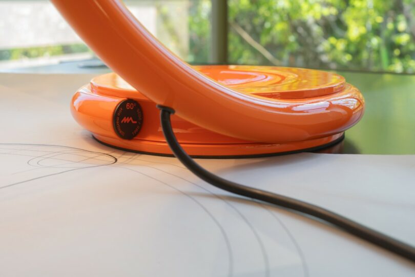 Close-up of the orange Serpente lamp base resting on a desk with architectural technical drawings.