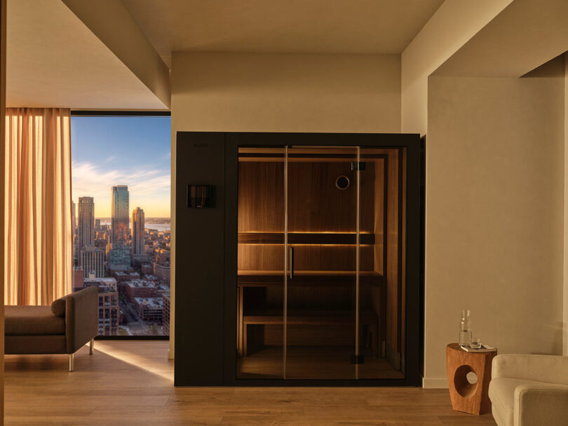 A minimalist indoor sauna with glass doors and a city view at dusk.