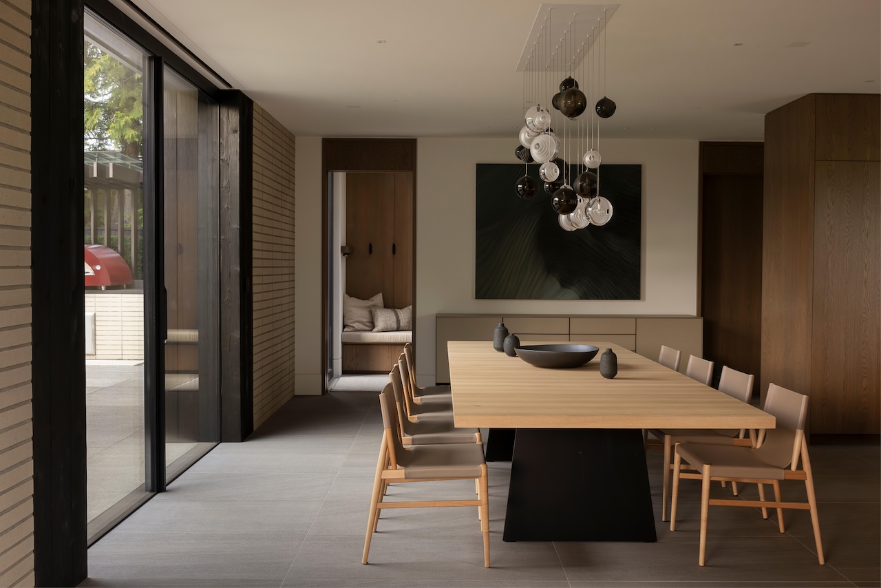 Minimalist dining room with a long wooden table, abstract wall art, and glass doors leading to a patio.
