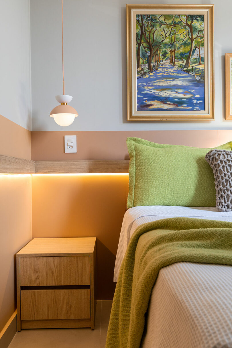 A bedroom corner featuring a wooden nightstand, green bedding, and a framed painting under soft pendant lighting.