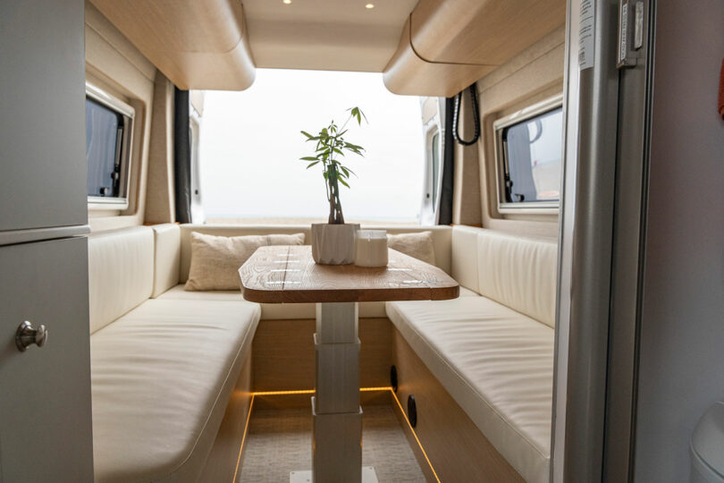 The U-shaped lounge area of a camper van with a wooden table, white cushions, and ambient lighting.