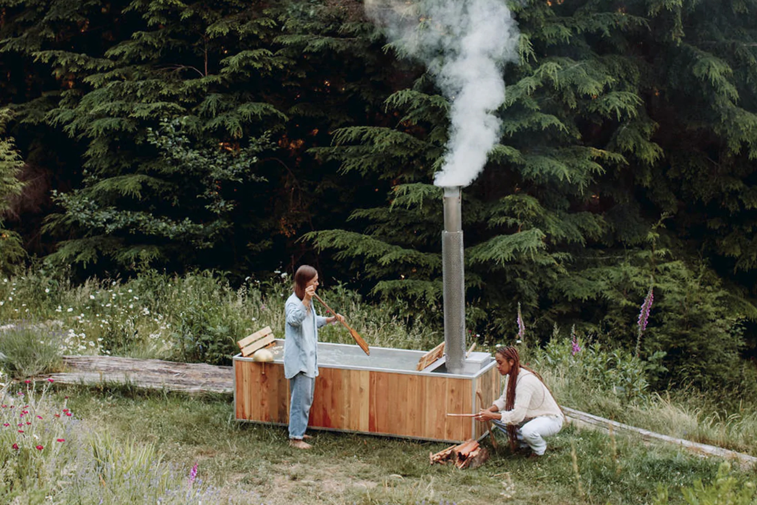Two people enjoying a rustic wooden hot tub in a natural woodland environment.