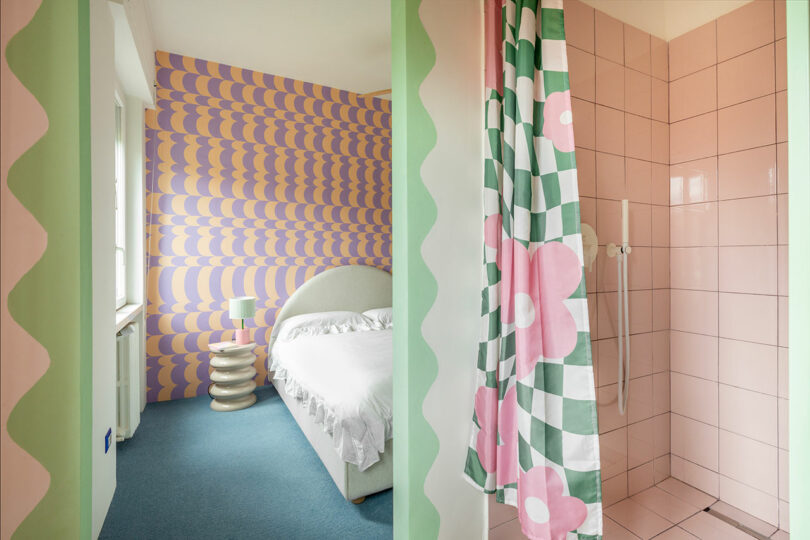 View from a wavy-bordered bedroom into a pink-tiled bathroom with a floral curtain.