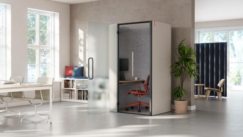 A glass-walled soundproof work pod featuring a red ergonomic chair and minimalist desk in a bright office.