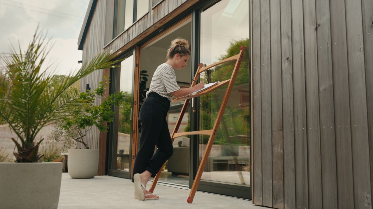 A woman sketching at a wooden desk on a modern wooden patio surrounded by plants.