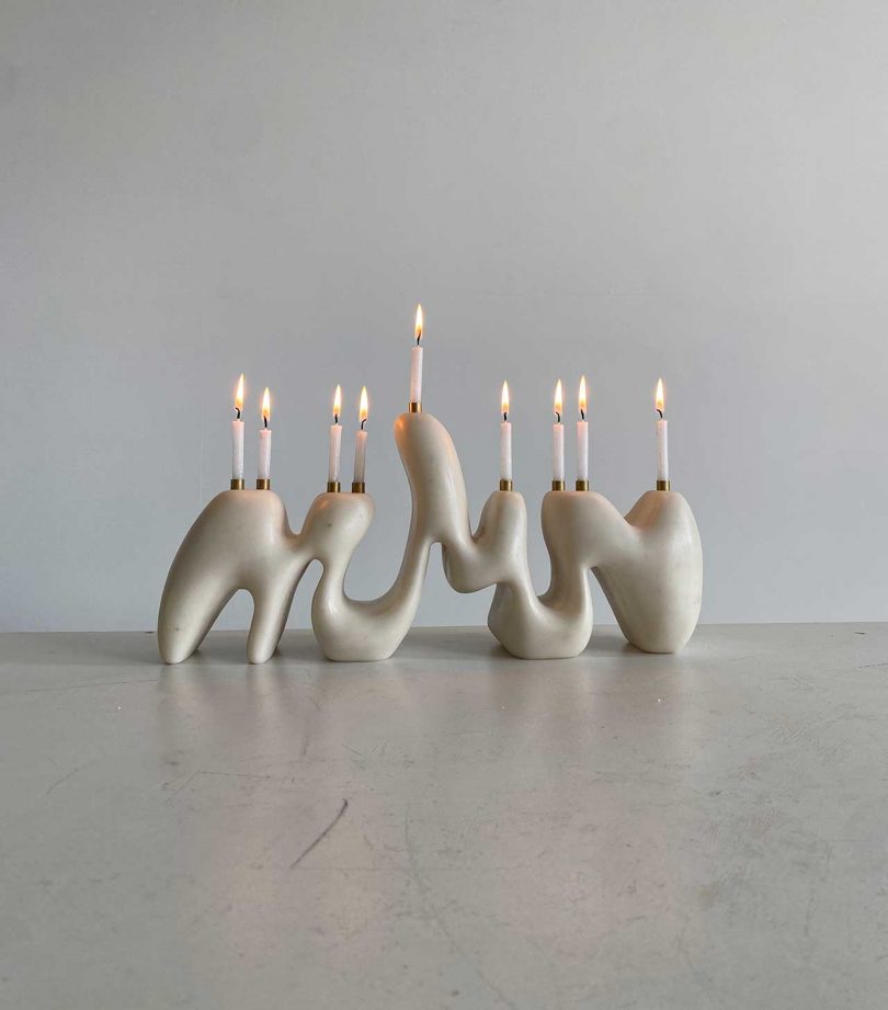 An off-white modern curvy organic menorah with white candles lit against a neutral background.