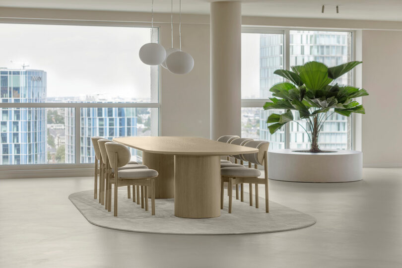 Modern minimalist dining room with a light wood oval table and floor-to-ceiling windows showing a city view.